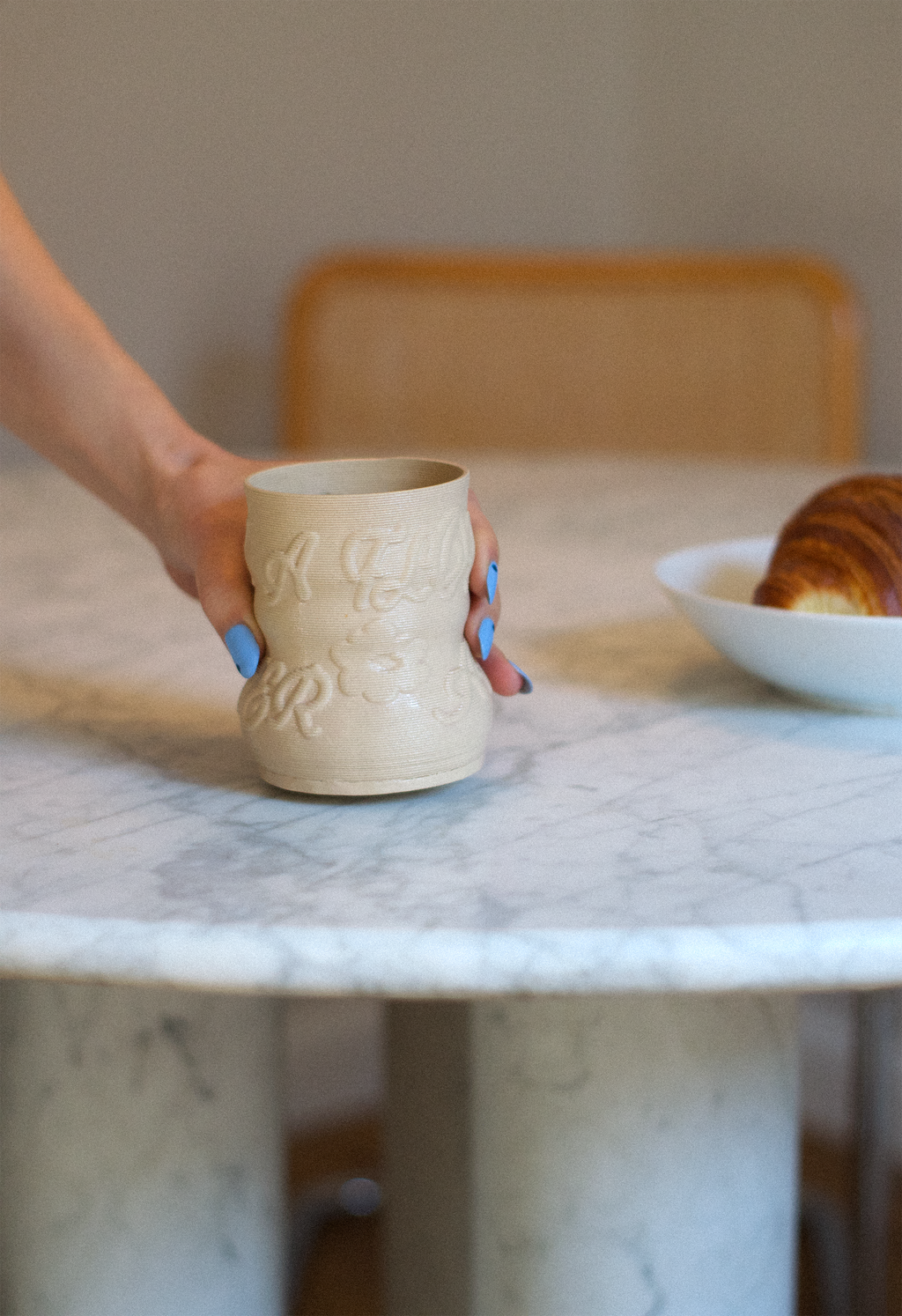 Lifestyle shot of a hand holding a cashew-colored 3D-printed clay matcha cup with a raised calligraphy pattern by GLINA3, resting on a marble table.