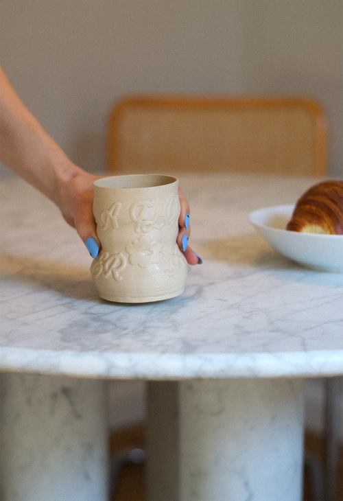 Lifestyle shot of a hand holding a cashew-colored 3D-printed clay matcha cup with a raised calligraphy pattern by GLINA3, resting on a marble table.