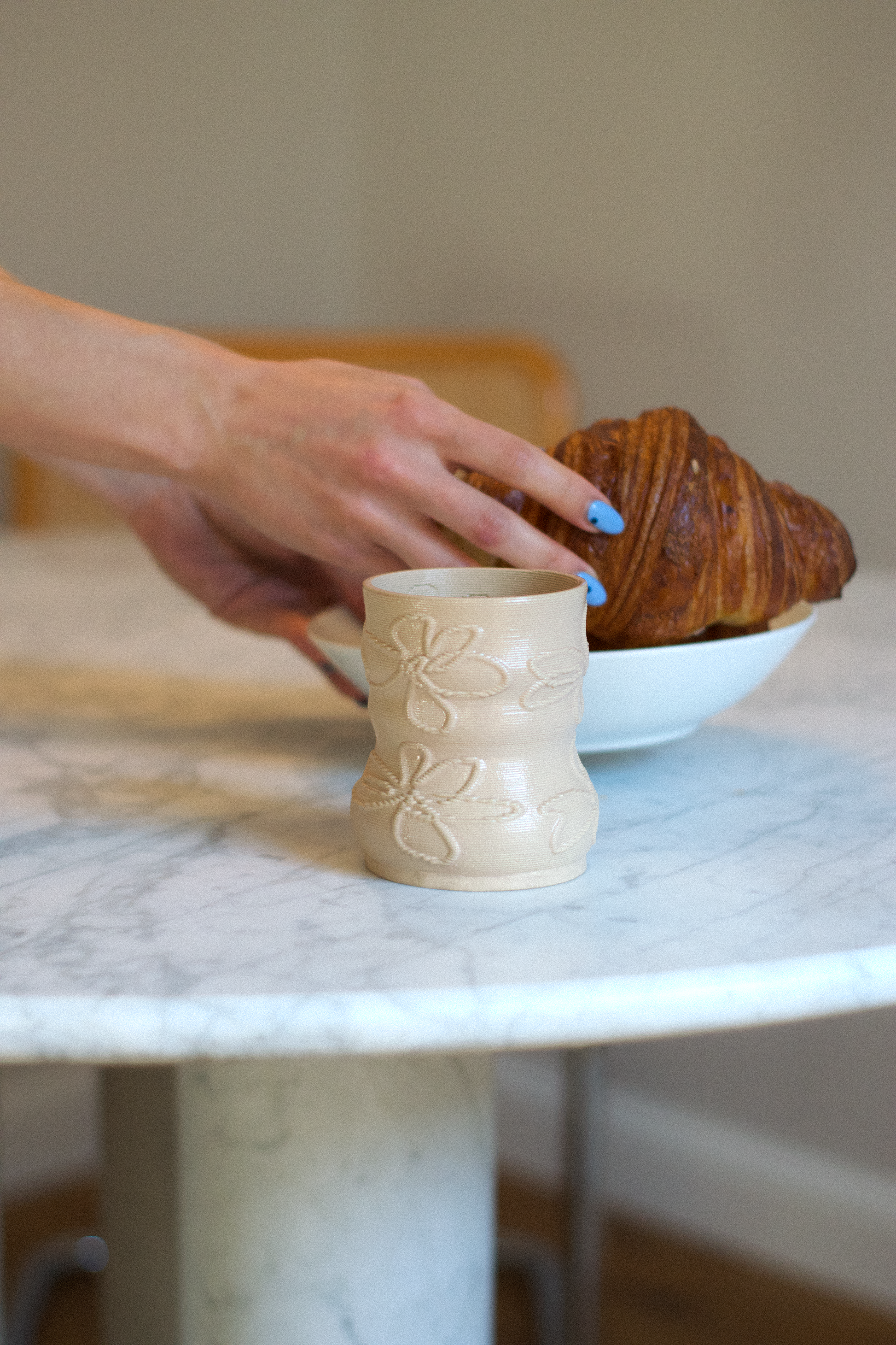 Lifestyle shot of a cashew-colored 3D-printed clay matcha cup by GLINA3, resting on a marble table with a croissant in the background.
