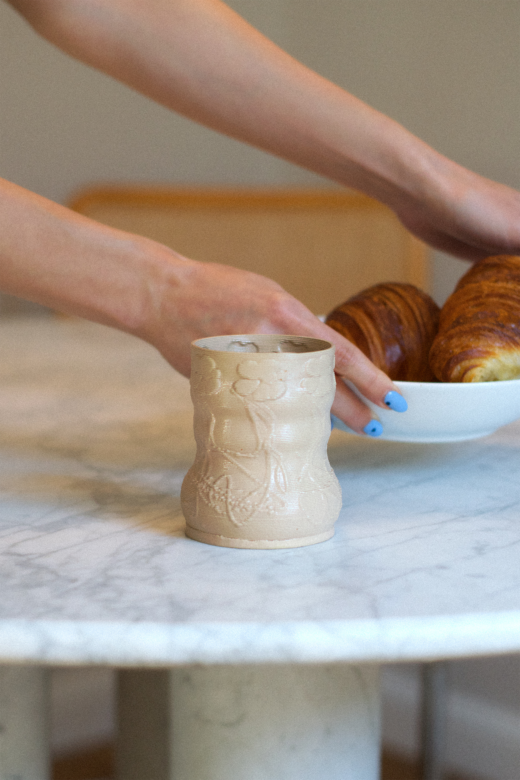 Lifestyle shot of a cashew-colored 3D-printed clay matcha cup with a rooted flower pattern by GLINA3, resting on a marble table next to a bowl of croissants.