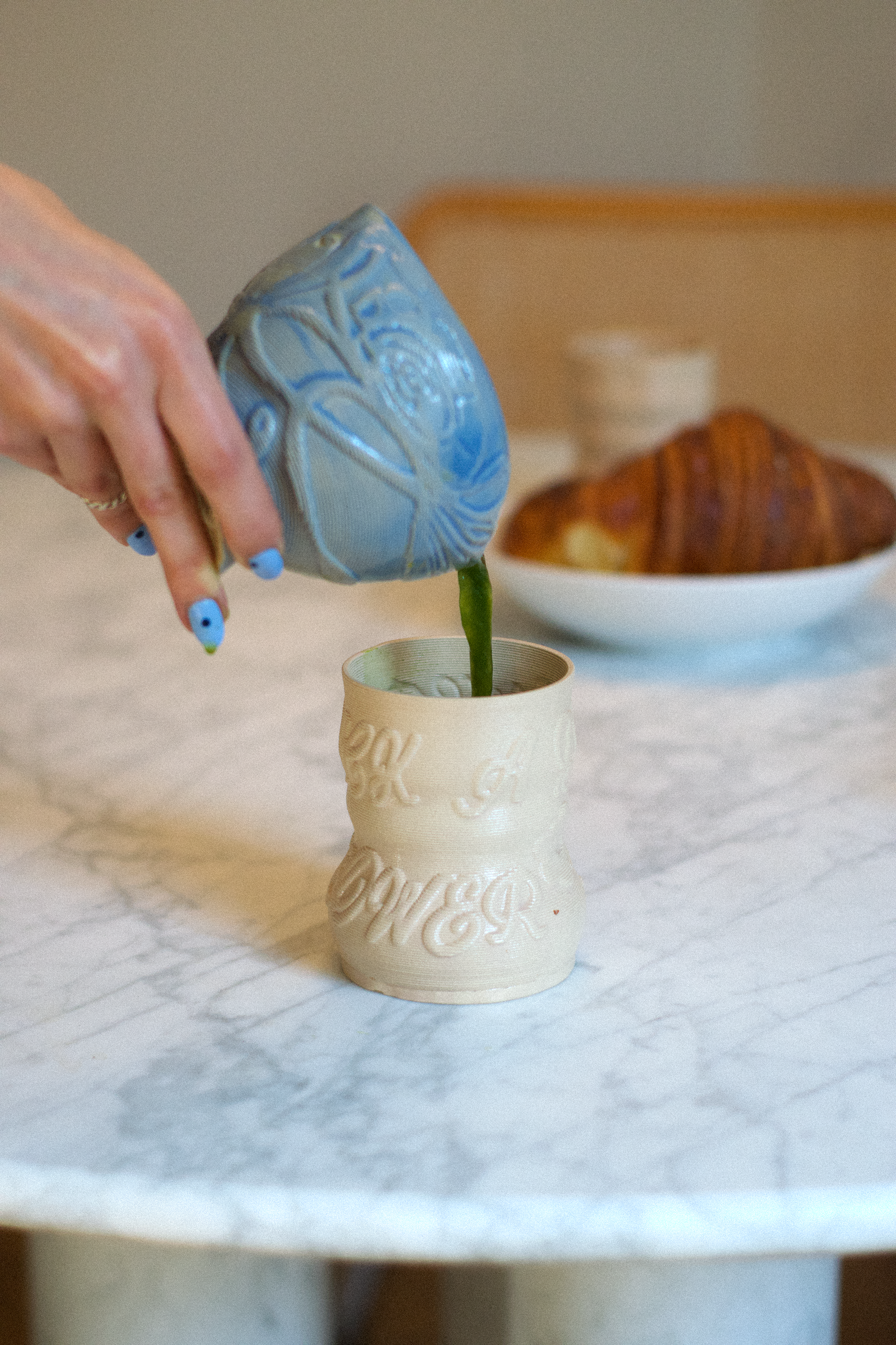Lifestyle shot of a hand pouring green matcha from a blue 3D-printed clay ceremonial bowl into a cashew-colored matcha cup by GLINA3, set on a marble table.
