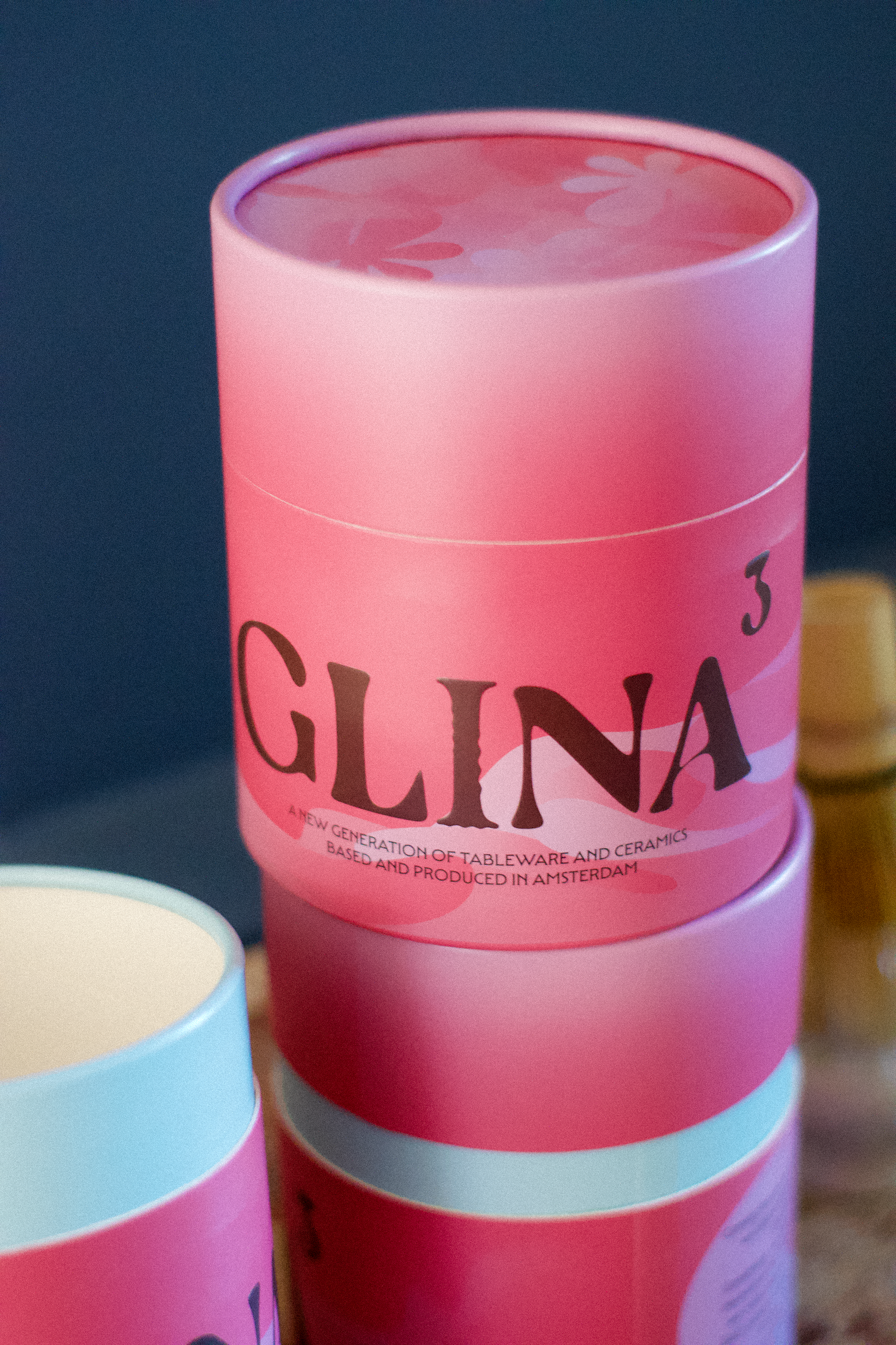 Close-up of the branded pink cylindrical packaging for GLINA3, featuring the text "A new generation of tableware and ceramics, based and produced in Amsterdam.